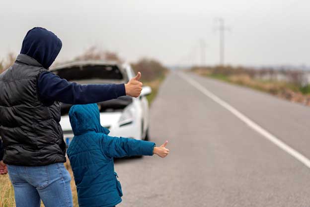 Father and child standing on the side of the road with their thumbs extended, waiting for a ride. Their white, broken-down car parked behind them with its hood up